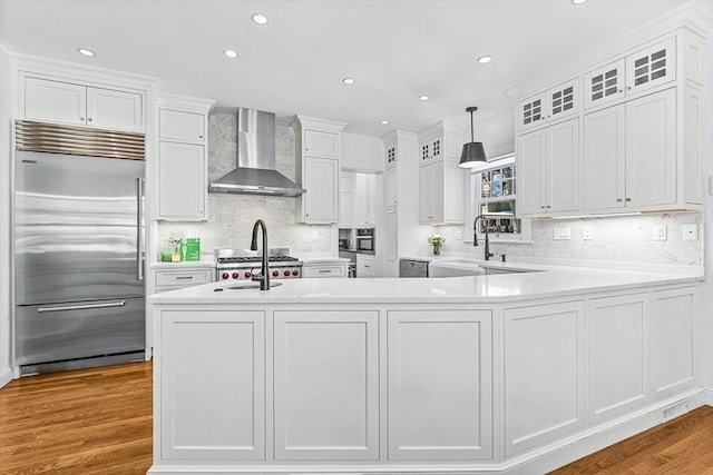 kitchen featuring decorative light fixtures, light countertops, glass insert cabinets, built in refrigerator, and wall chimney exhaust hood