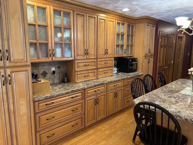 kitchen featuring light wood finished floors, brown cabinetry, stone counters, backsplash, and glass insert cabinets