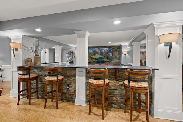 bar with a decorative wall, bar area, light wood-style floors, and ornate columns