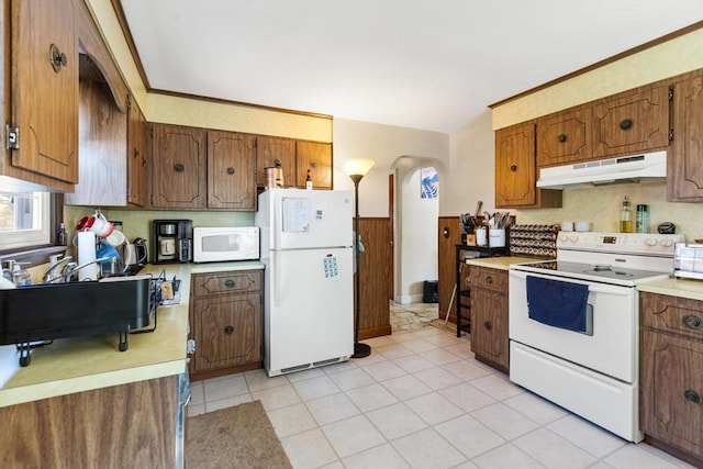 kitchen featuring brown cabinetry, wainscoting, light countertops, under cabinet range hood, and white appliances