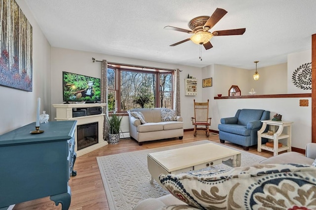 living room featuring a ceiling fan, baseboards, light wood-style flooring, a textured ceiling, and a glass covered fireplace