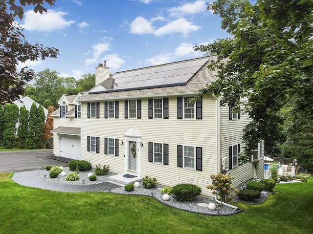 colonial house featuring a chimney, a front lawn, a garage, aphalt driveway, and roof mounted solar panels