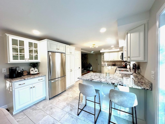 kitchen with freestanding refrigerator, a peninsula, a breakfast bar area, glass insert cabinets, and light stone countertops