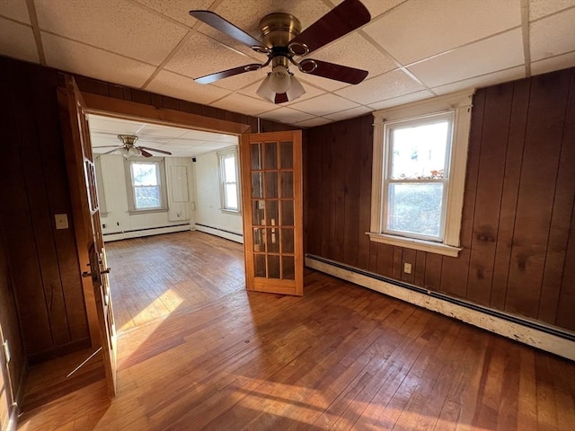 unfurnished room featuring wood walls, baseboard heating, a drop ceiling, and hardwood / wood-style flooring