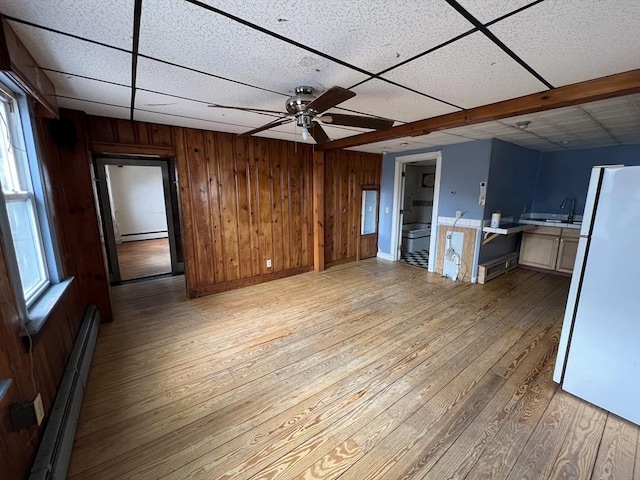 unfurnished living room featuring light wood-style flooring, wooden walls, baseboard heating, and a drop ceiling