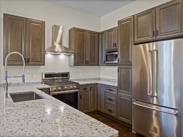 kitchen with stainless steel appliances, a sink, light stone countertops, wall chimney exhaust hood, and dark brown cabinetry