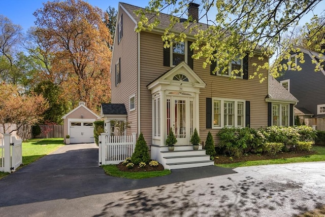 colonial house with driveway, a chimney, a detached garage, fence, and a shingled roof