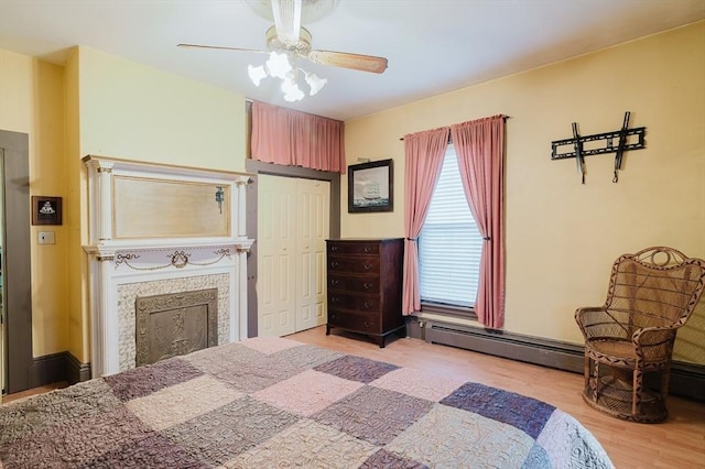 bedroom with light wood-type flooring, a closet, a fireplace, a baseboard radiator, and a ceiling fan