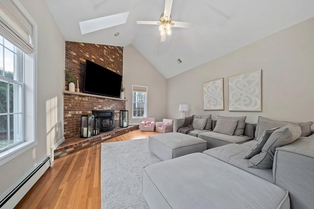 living room featuring hardwood / wood-style flooring, ceiling fan, a wood stove, baseboard heating, and vaulted ceiling with skylight