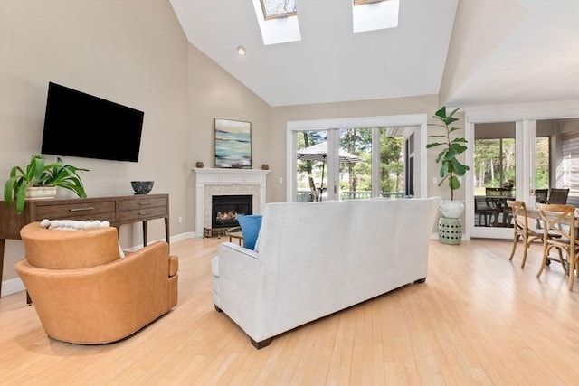 living area featuring a warm lit fireplace, high vaulted ceiling, baseboards, light wood-type flooring, and a skylight