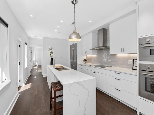 kitchen featuring wall chimney exhaust hood, sink, decorative light fixtures, white cabinetry, and a center island with sink