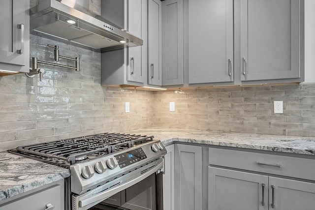 kitchen with gray cabinets, stainless steel gas stove, decorative backsplash, light stone countertops, and wall chimney exhaust hood