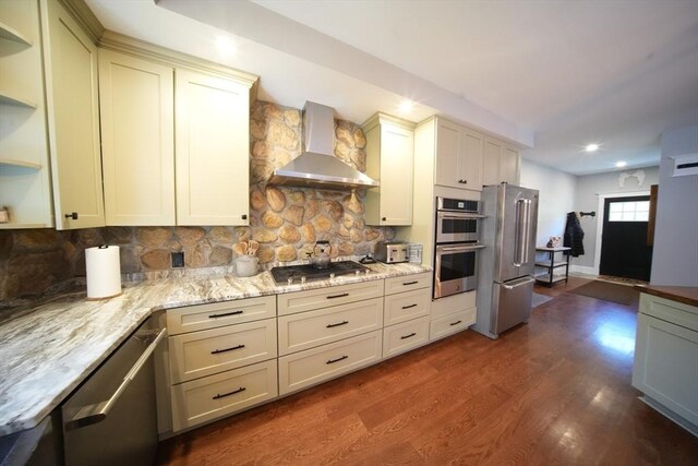 kitchen with appliances with stainless steel finishes, light stone counters, wall chimney range hood, dark wood-type flooring, and tasteful backsplash