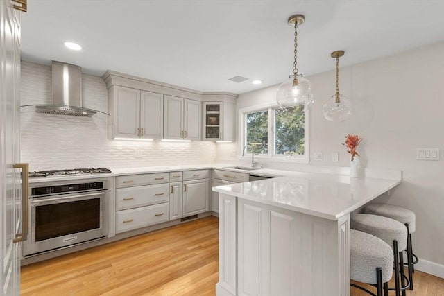 kitchen featuring a breakfast bar area, stainless steel appliances, a peninsula, wall chimney exhaust hood, and light wood finished floors