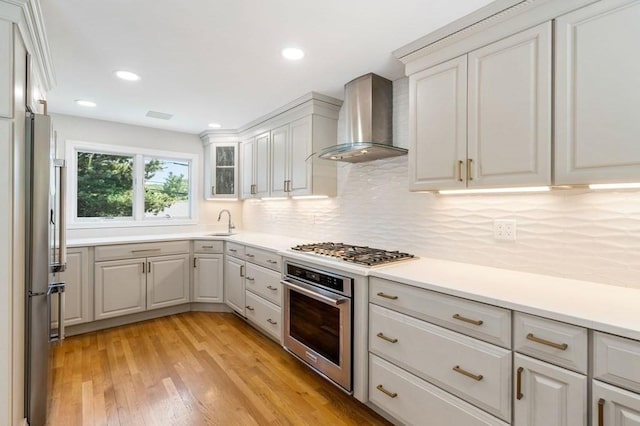 kitchen featuring stainless steel appliances, a sink, wall chimney range hood, light wood finished floors, and tasteful backsplash