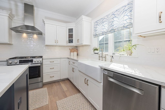 kitchen with appliances with stainless steel finishes, wall chimney range hood, white cabinetry, and light hardwood / wood-style flooring