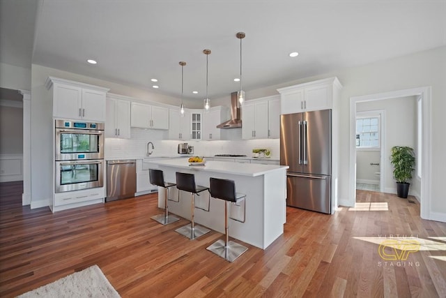 kitchen with wall chimney range hood, white cabinetry, hanging light fixtures, stainless steel appliances, and a kitchen island