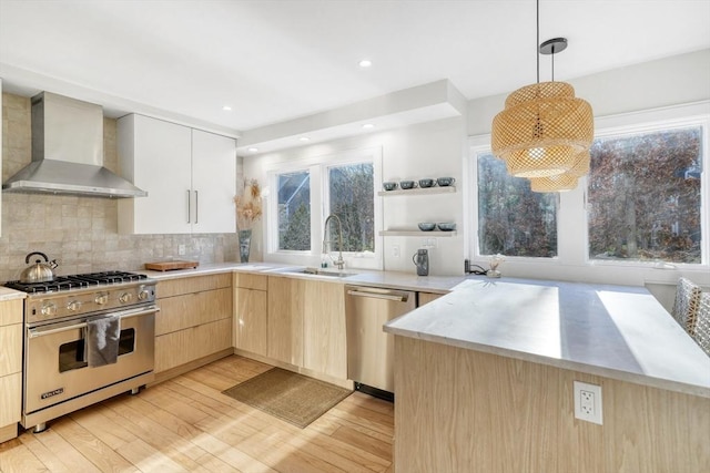 kitchen featuring appliances with stainless steel finishes, light brown cabinetry, wall chimney range hood, decorative light fixtures, and light hardwood / wood-style flooring