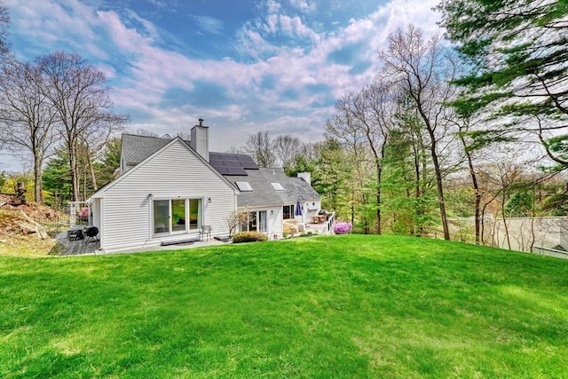 rear view of property with a lawn, a chimney, roof mounted solar panels, and a patio area