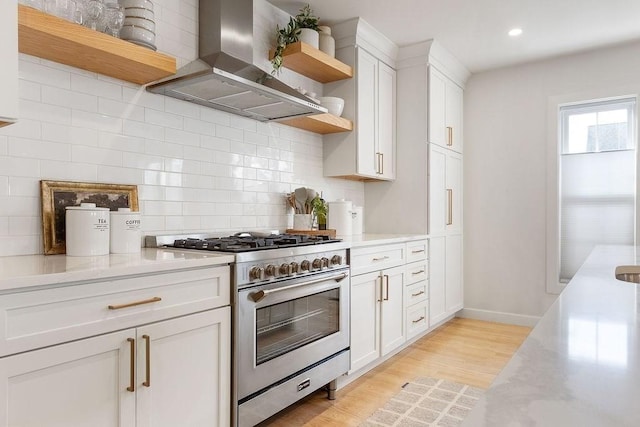 kitchen with high end range, light stone counters, wall chimney range hood, white cabinets, and light hardwood / wood-style floors