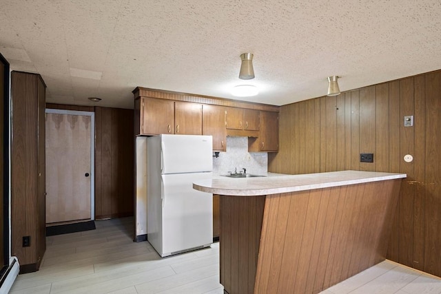 kitchen featuring freestanding refrigerator, wooden walls, a peninsula, brown cabinets, and light countertops