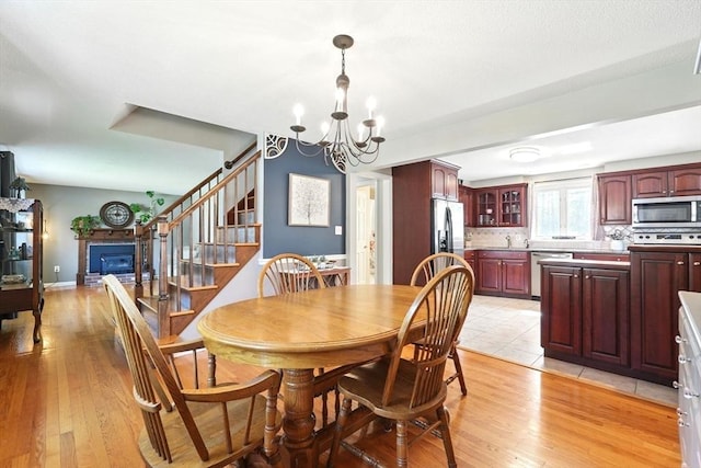 dining space featuring light wood-type flooring, stairway, baseboards, a notable chandelier, and a glass covered fireplace