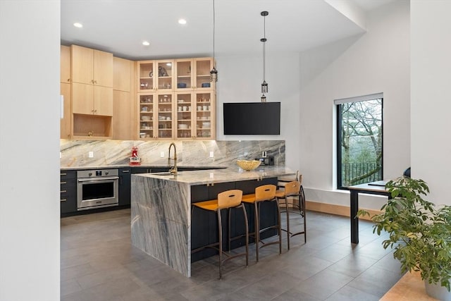 kitchen with stainless steel oven, light brown cabinetry, backsplash, light stone counters, and recessed lighting