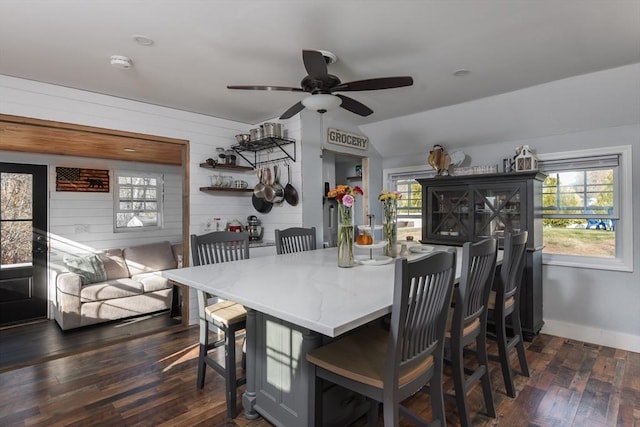 dining area featuring wooden walls, dark wood-type flooring, a ceiling fan, and baseboards