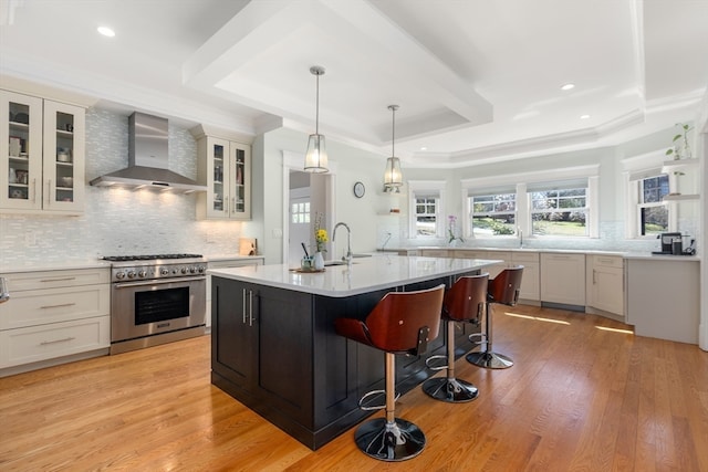 kitchen with light wood-type flooring, a center island with sink, wall chimney exhaust hood, and stainless steel stove