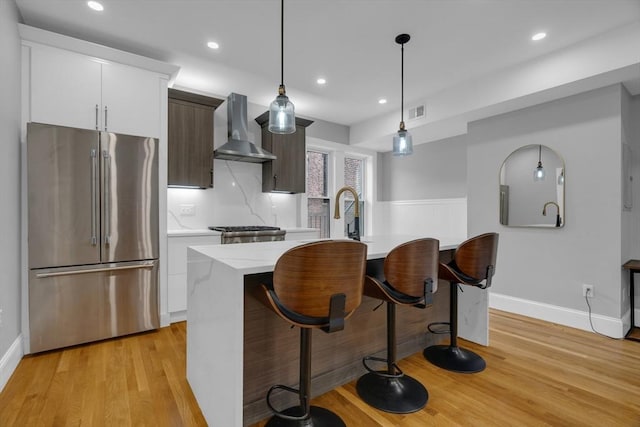 kitchen featuring pendant lighting, range, wall chimney range hood, a kitchen island with sink, and high quality fridge