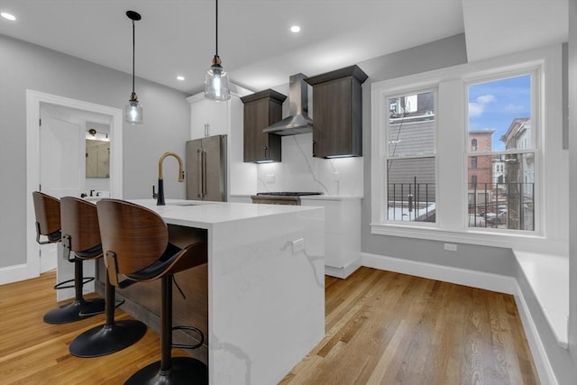 kitchen with decorative light fixtures, an island with sink, wall chimney range hood, high quality fridge, and dark brown cabinetry