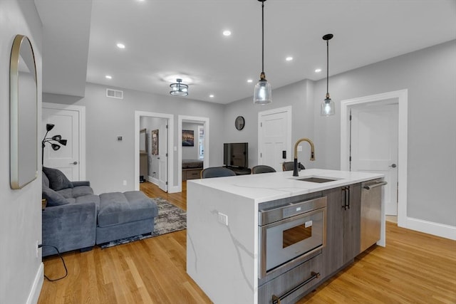 kitchen with sink, light hardwood / wood-style floors, an island with sink, stainless steel dishwasher, and hanging light fixtures