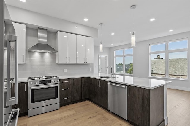 kitchen with wall chimney range hood, light wood-style flooring, stainless steel appliances, white cabinetry, and a sink