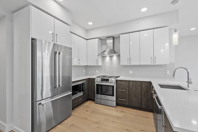 kitchen with a sink, light countertops, appliances with stainless steel finishes, wall chimney range hood, and light wood-type flooring