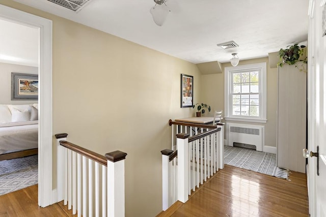 corridor featuring wood finished floors, radiator heating unit, visible vents, and an upstairs landing