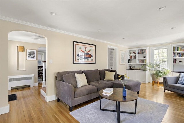living area featuring light wood-style floors, arched walkways, radiator heating unit, baseboards, and ornamental molding