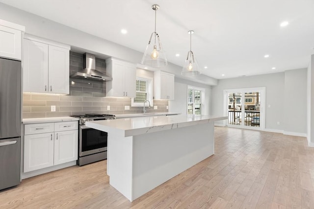 kitchen with wall chimney exhaust hood, a kitchen island, appliances with stainless steel finishes, light wood-style flooring, and backsplash