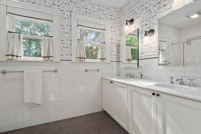 bathroom featuring tile walls, double vanity, a tile shower, a wainscoted wall, and wallpapered walls