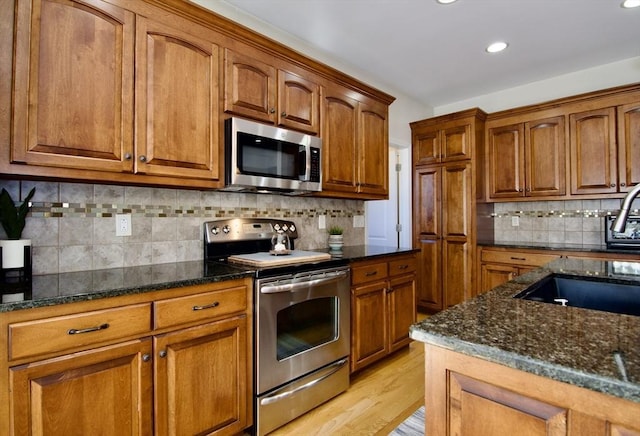 kitchen with dark stone countertops, brown cabinetry, a sink, light wood-style floors, and appliances with stainless steel finishes