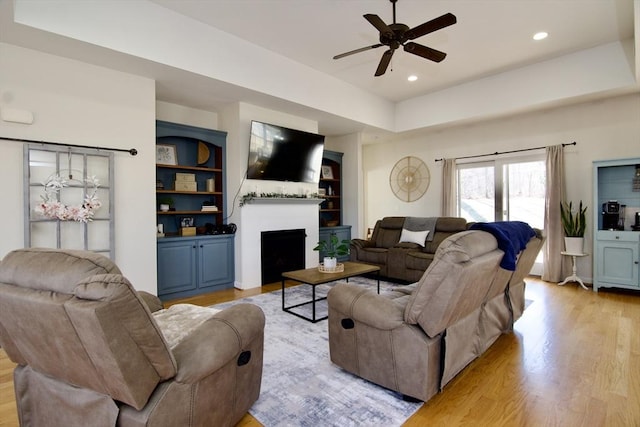 living area featuring recessed lighting, a fireplace, ceiling fan, a raised ceiling, and light wood-type flooring