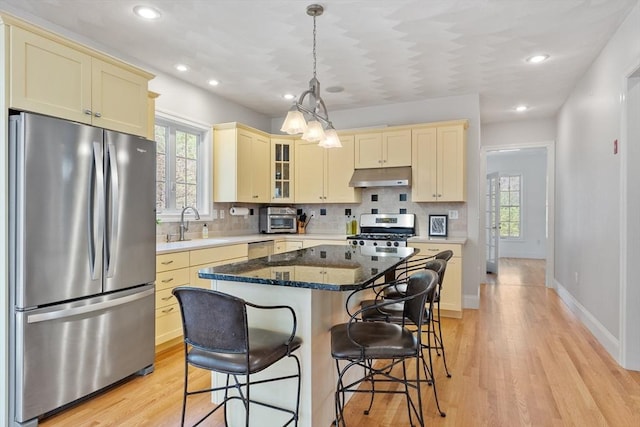 kitchen with stainless steel appliances, under cabinet range hood, cream cabinets, a sink, and light wood-style floors