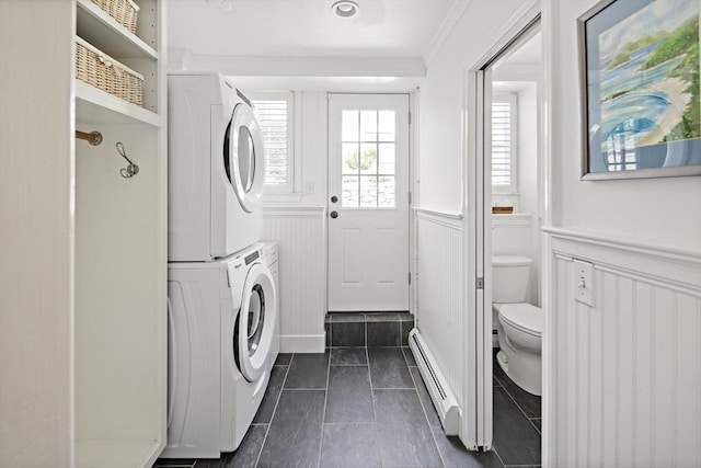 laundry room featuring baseboard heating, stacked washer / drying machine, wainscoting, dark tile patterned flooring, and ornamental molding
