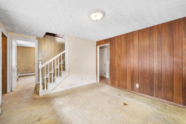 carpeted empty room with a textured ceiling, wooden walls, baseboards, stairs, and baseboard heating