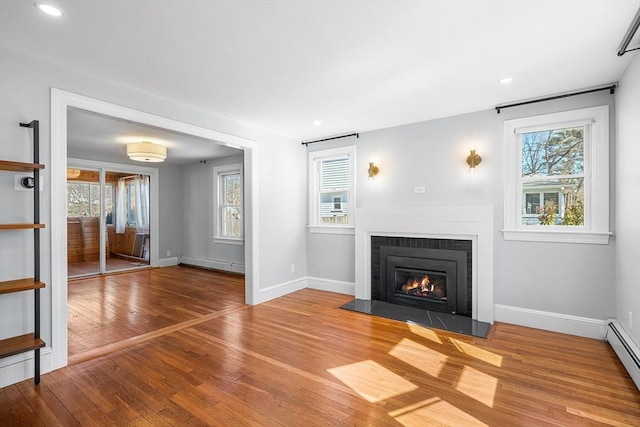 unfurnished living room with wood-type flooring, baseboard heating, a healthy amount of sunlight, and a glass covered fireplace