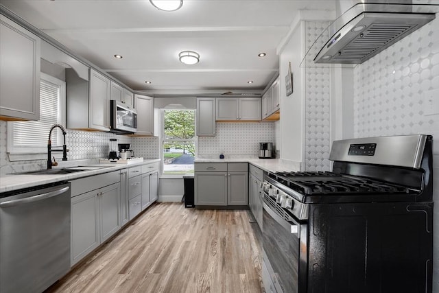 kitchen with stainless steel appliances, gray cabinets, a sink, wall chimney exhaust hood, and light wood-type flooring