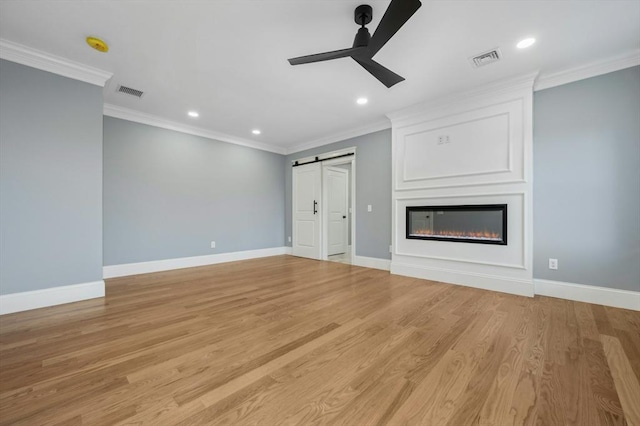 unfurnished living room with ceiling fan, a large fireplace, a barn door, light hardwood / wood-style flooring, and ornamental molding