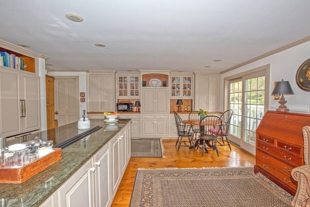 kitchen featuring white cabinets, light wood-style floors, dark stone counters, ornamental molding, and glass insert cabinets
