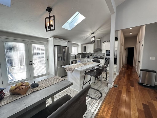 kitchen featuring white cabinetry, wall chimney exhaust hood, vaulted ceiling with skylight, stainless steel refrigerator with ice dispenser, and pendant lighting