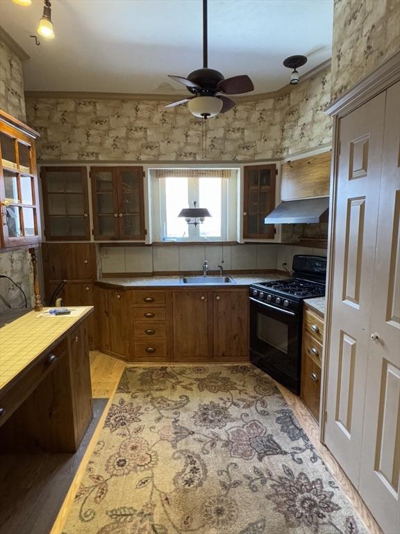 kitchen with glass insert cabinets, gas stove, light wood-style floors, a sink, and wall chimney range hood