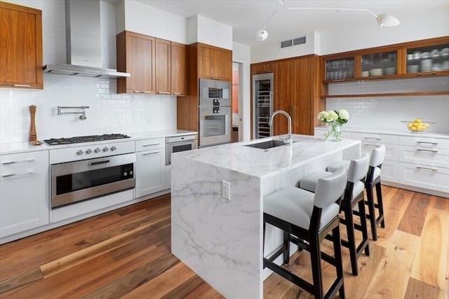 kitchen featuring wood-type flooring, wall chimney exhaust hood, white cabinets, a kitchen island with sink, and sink